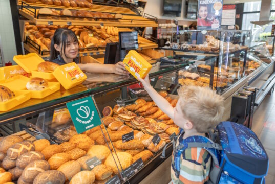 Evertzberg-Mitarbeiterin Dajana Murati übergibt Erstklässler David eine gefüllte, gelbe Snackbox. (Foto: © Thomas Wunsch)