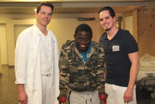 Nach überstandener Operation kämpft sich Steven Jacob Amunga (mi.) Schritt für Schritt in ein Leben ohne Behinderung. An seiner Seite: Dr. Oliver Voß (li.), Oberarzt der Klinik für Unfallchirurgie, Orthopädie und Handchirurgie, und Physiotherapeut Michele Lorenzo Patricelli. (Foto: © Bastian Glumm)