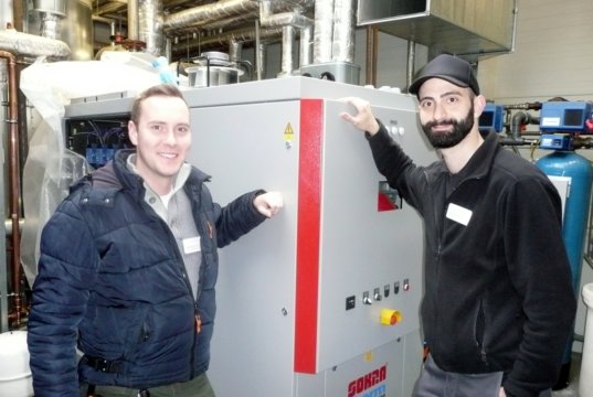 Patrick Preiss (li.), Energiemanager der Technischen Abteilung der Kplus Gruppe, und Kemal Hilmi Dogan, Technischer Betriebsleiter der Genesis Zentralküche an der Alsenstraßen, vor dem 2017 dort installierten neuen Blockheizkraftwerk. (Foto: © Kplus Gruppe)