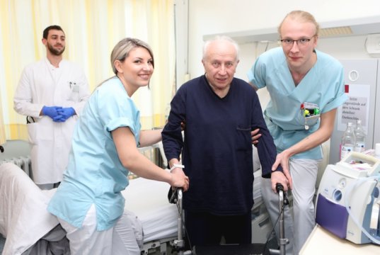 Hermine Harder (li.) und Philipp Röltgen (re.) helfen einem Patienten der St. Lukas Klinik. Im Hintergrund Assistenzarzt Tariq Al-Hindi. (Foto: © Kplus Gruppe)