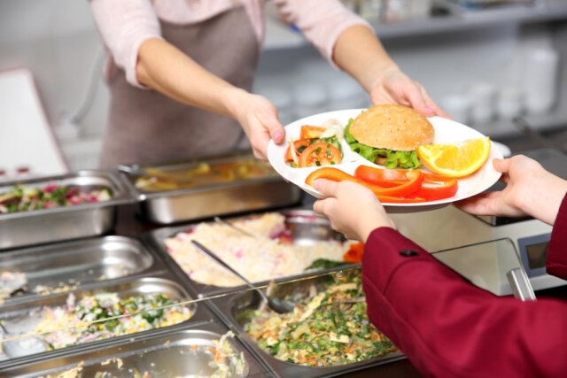 Pleasant woman giving lunch to school girl in cafeteria Die Stadt Solingen organisiert derzeit eine neue Lösung für die Mittagsverpflegung an der Alexander-Coppel-Gesamtschule und an der Geschwister-Scholl-Schule. Sie stellt den Mensabetrieb an beiden Schulen neu auf. (Symbolfoto: © Africa Studio / Adobe Stock)