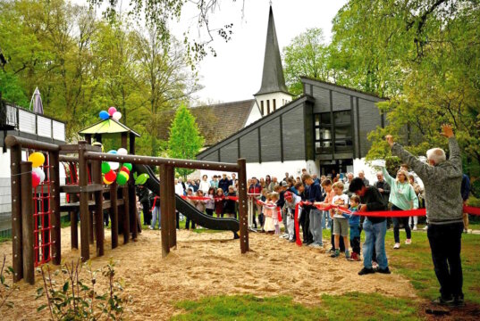 Kinder durchschneiden das rote Band zur Eröffnung des neuen Spielplatz hinter dem Aufderhöher Gemeindezentrum, zahlreiche Besucher verfolgen den offiziellen Start. (Foto: © Petra Schelkes)