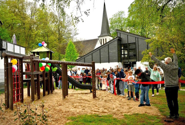 Kinder durchschneiden das rote Band zur Eröffnung des neuen Spielplatz hinter dem Aufderhöher Gemeindezentrum, zahlreiche Besucher verfolgen den offiziellen Start. (Foto: © Petra Schelkes)