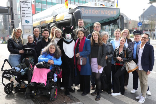 Teilnehmer und Projektbeteiligte bei der Auftaktfahrt der neuen O-Bus-Quiztour am Graf-Wilhelm-Platz: mit dabei Kater Salabim (im Kostüm: Sandra Kratz), Schirmherrin Sonja Flemm (li. daneben), Bürgermeisterin Ioanna Zacharaki (re. neben dem Kater) sowie Sandra-Janine Schneider (7. v. rechts) vor dem Sonderwagen der Linie 681. (Foto: © Bastian Glumm)